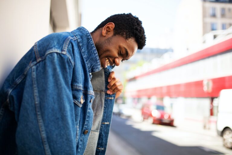 Side of smiling young black man leaning against wall and holding denim jacket outside